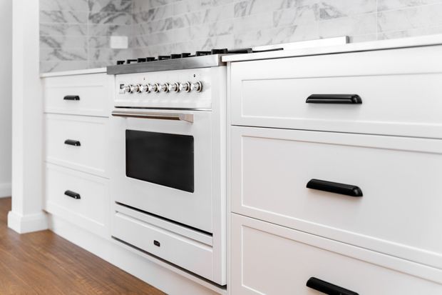 Close-up view of a bright white fitted kitchen featuring a freestanding-style range cooker integrated between Shaker-style drawer and cabinet units. The cabinetry has raised-panel fronts with black bar handles, a white benchtop, and a marble-look tiled splashback. Warm timber flooring contrasts with the predominantly white and black palette.