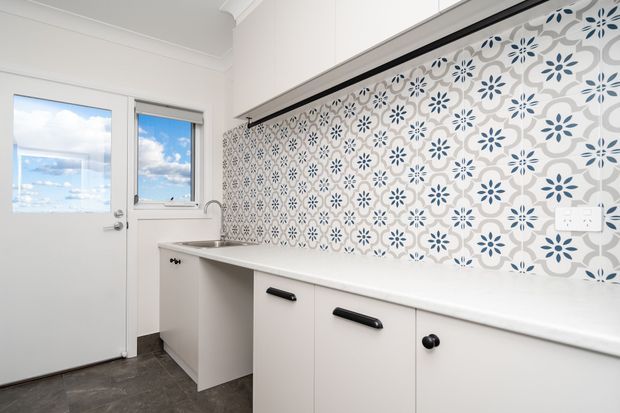 A compact, bright laundry room with white built-in cabinetry and benchtops, a deep utility sink, and a patterned blue-and-white tiled splashback. The room has a glazed exterior door and window, dark flooring, and simple contemporary fittings.