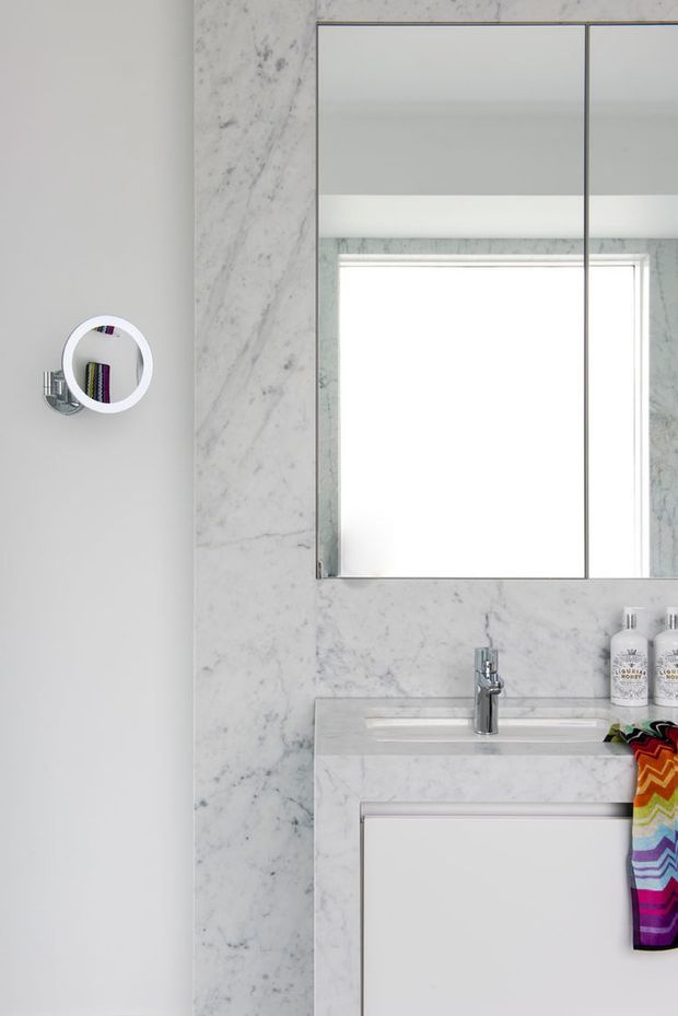 A compact, bright bathroom vanity with a white marble-clad wall and benchtop, integrated rectangular basin, chrome single-lever faucet, mirrored wall cabinet, and a wall-mounted circular magnifying mirror. White cabinetry and marble create a clean, refined look, with colourful towels adding a small accent.