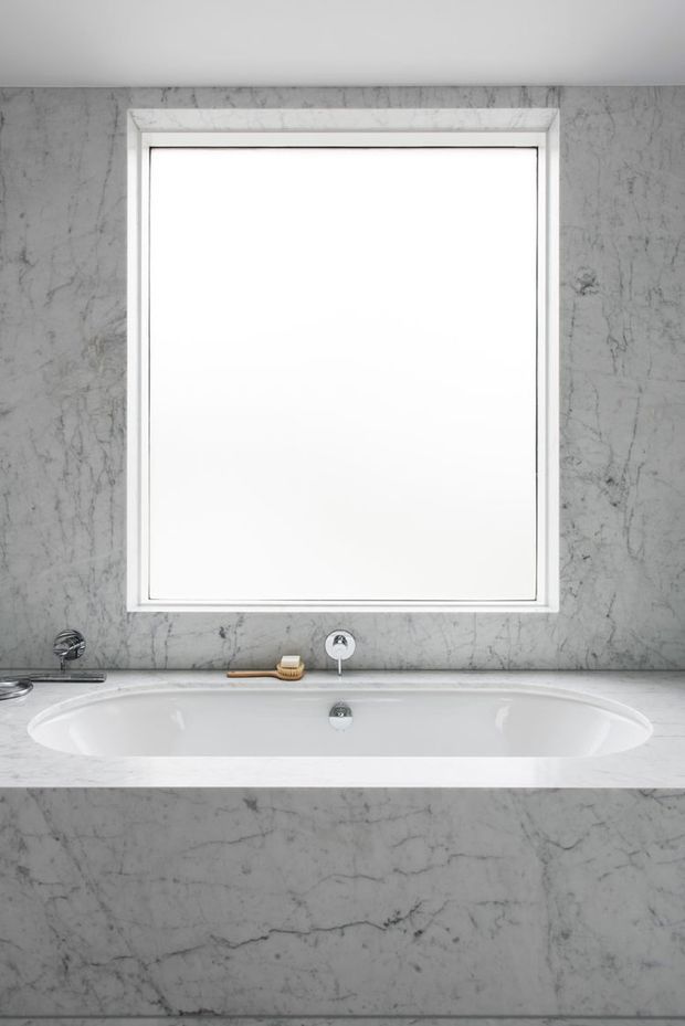 Minimalist bathroom vignette featuring a built-in white bathtub set into a grey veined stone surround, with a matching stone wall and window frame. A simple wall-mounted faucet and hand shower are visible, creating a clean monochromatic composition.
