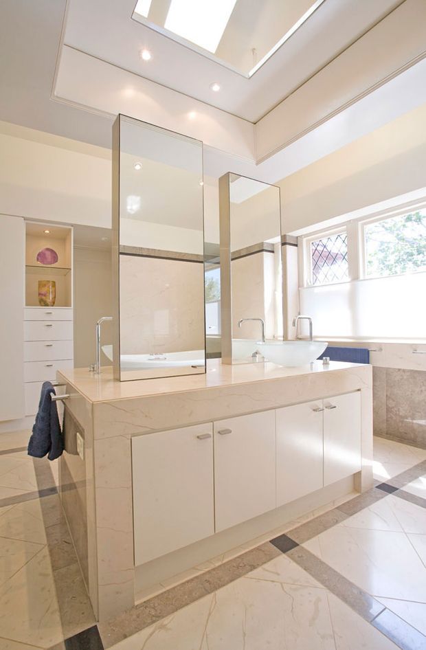 Bright, spacious bathroom with a central double vanity island featuring a pale stone benchtop, integrated cabinetry, twin vessel basins and tall mirrors. The room includes marble-look tiled flooring and wall finishes, a bathtub visible behind the vanity, built-in shelving, high windows with diamond-pattern glazing, recessed lighting and a large skylight.
