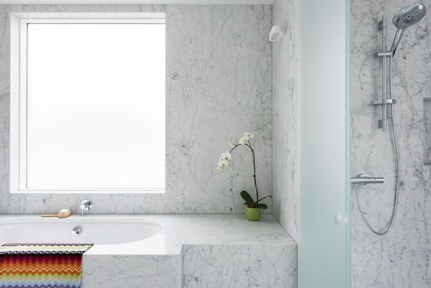 Bright contemporary bathroom featuring a white bathtub, marble-look wall and bench surfaces, a frameless shower enclosure with chrome shower fittings, and a small orchid. A colourful patterned textile adds an accent at the bath.