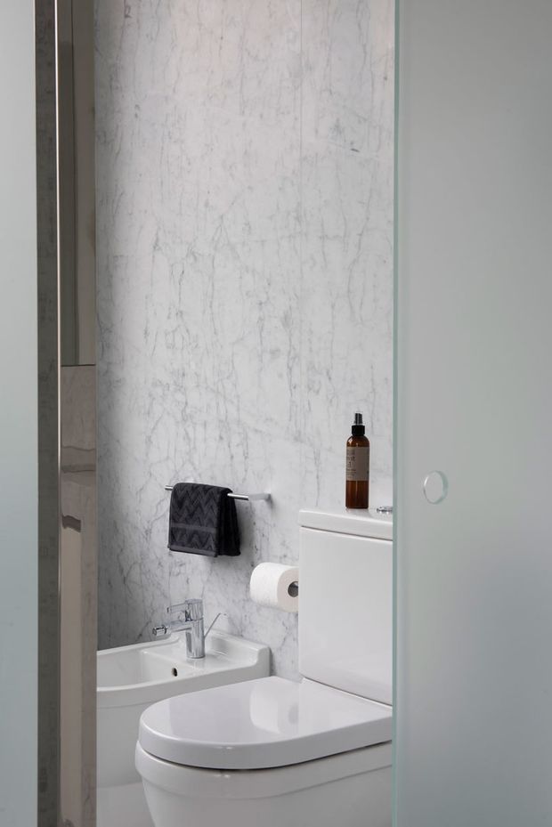 Compact contemporary bathroom viewed through a partially open glass door, featuring a white toilet, small white basin with chrome tapware, wall-mounted towel rail, toilet roll holder, and a full-height white marble-look wall surface with subtle grey veining. The palette is clean and neutral with grey framing and frosted or translucent glass.