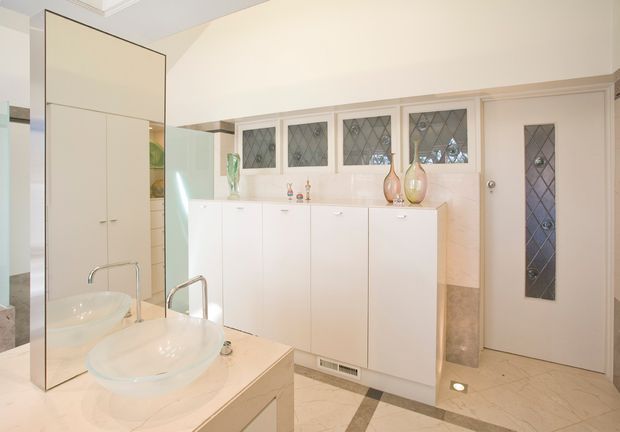 Bright, refined bathroom with two vessel basins on a pale stone vanity, extensive white built-in cabinetry, mirrored surfaces, decorative diamond-leaded windows, and a matching leaded-glass door. The floor uses pale stone tiles with darker geometric border inlays, creating a classic high-end feel.