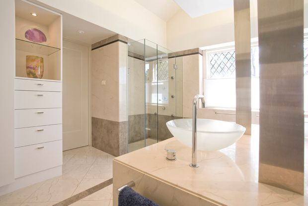 Bright, spacious luxury bathroom featuring a freestanding glass-enclosed shower, a sculptural white vessel basin on a pale stone vanity, polished chrome tapware, marble-look wall and floor finishes, built-in white storage, and diamond-patterned windows. The room has a refined traditional feel with strong natural light and reflective surfaces.