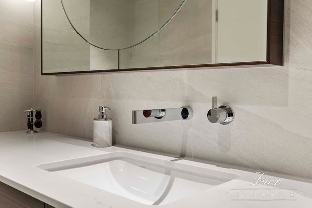 Close-up of a refined bathroom vanity with a white integrated basin, wall-mounted chrome tapware and controls, pale stone or porcelain wall finish, and a large timber-framed mirror. The restrained palette and clean geometric fixtures create a polished, contemporary feel.