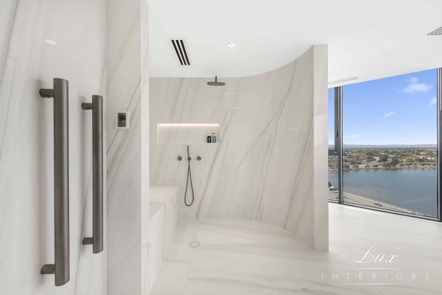 A bright, luxurious contemporary bathroom featuring a curving walk-in shower enclosed by large white marble-look panels, dark brushed-metal shower fittings, recessed shelf lighting, and frameless glazing. Through the glazing, a waterfront urban view is visible.