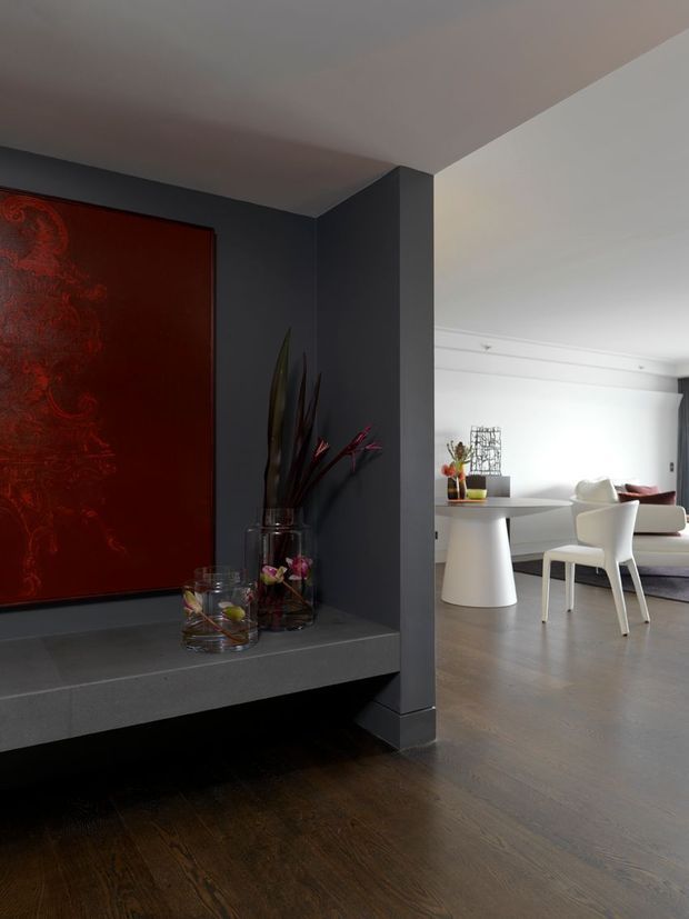 Interior view of a refined contemporary home, looking past a dark charcoal feature wall with a large red artwork and built-in floating ledge toward a bright dining and living area. The adjoining space contains a white pedestal dining table, white dining chairs, and a pale sofa. Dark timber flooring contrasts with the light furnishings and white ceiling, while decorative vases and sculptural branches sit on the ledge.