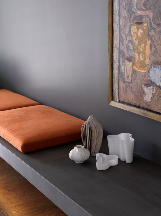 Close-up interior vignette of a dark, minimalist built-in platform or bench against a charcoal-grey wall, topped with rust-orange cushions, sculptural ceramic vessels, and a tall ribbed vase. A framed abstract artwork hangs above, while warm timber flooring is visible below. The composition has a refined, contemporary feel with strong tonal contrast between the dark surfaces, orange upholstery, white ceramics, and artwork.