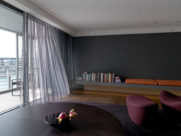 A refined contemporary living room with floor-to-ceiling glazing and sheer striped curtains opening to a balcony. The room features a dark charcoal feature wall, floating timber shelving with books and a long rust-coloured cushion, dark timber flooring, a large round black coffee table, and sculptural plum lounge chairs. The space has a restrained, moody palette with strong natural light from the glazing.