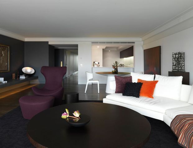 A contemporary open-plan living room with a large white sectional sofa, dark round coffee table, sculptural purple lounge chair, and dark feature cabinetry. The space opens toward a compact kitchen and dining area, with white walls, recessed ceiling details, timber flooring, and bold orange and purple cushions providing accents.