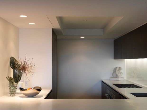 A compact contemporary kitchen viewed across a pale stone or solid-surface island. The room features dark wood or charcoal cabinetry, a white illuminated splashback, integrated cooktop, recessed ceiling lights, and a recessed ceiling detail. Minimal decorative objects sit on the island, including a glass vase and bowl.