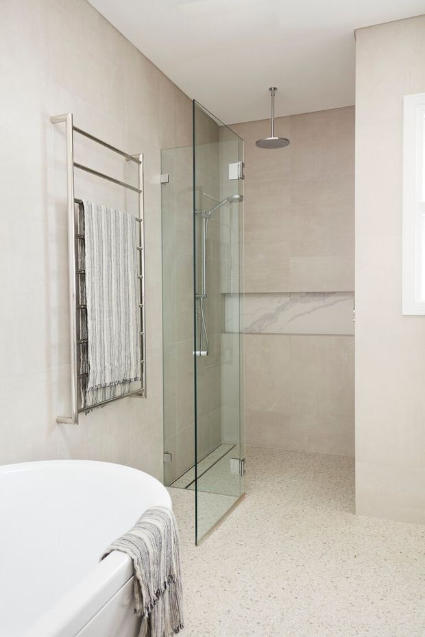 Bright contemporary bathroom featuring a freestanding white bathtub, frameless glass walk-in shower, wall-mounted stainless steel heated towel rail, large pale beige wall tiles, a marble-look horizontal feature band, and a light terrazzo-style floor. The room has a clean, refined, spa-like atmosphere with natural light from a side window.