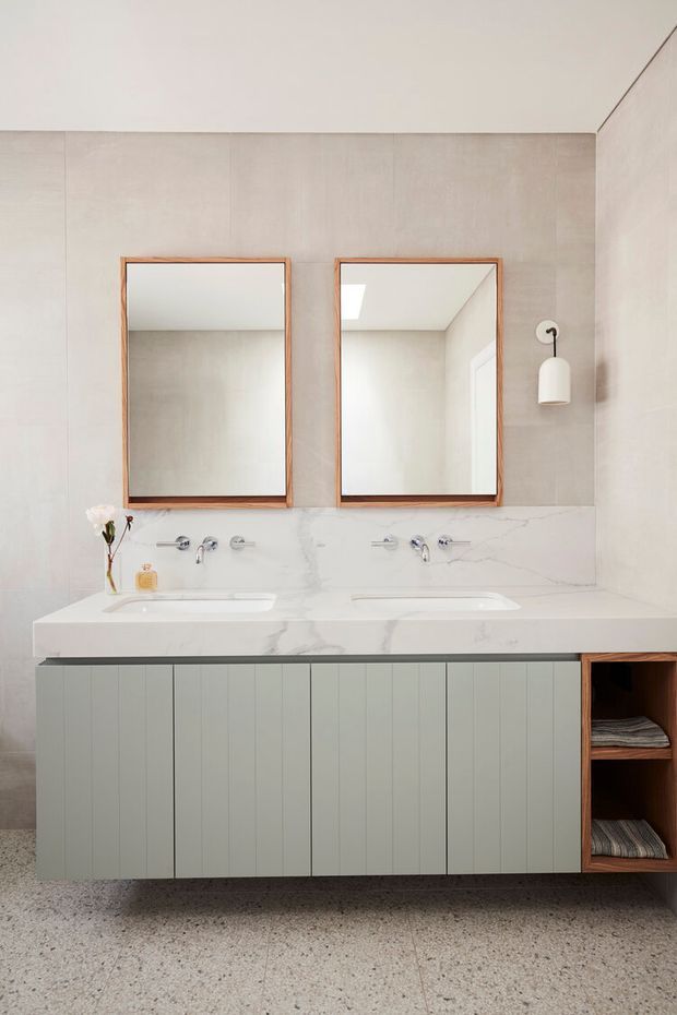 A bright contemporary bathroom featuring a double floating vanity with integrated white stone basins and marble-effect benchtop, pale vertically panelled cabinetry, open timber shelving, twin timber-framed mirrors, pale wall tiles, chrome tapware, and a terrazzo floor. A simple white wall sconce provides additional lighting.