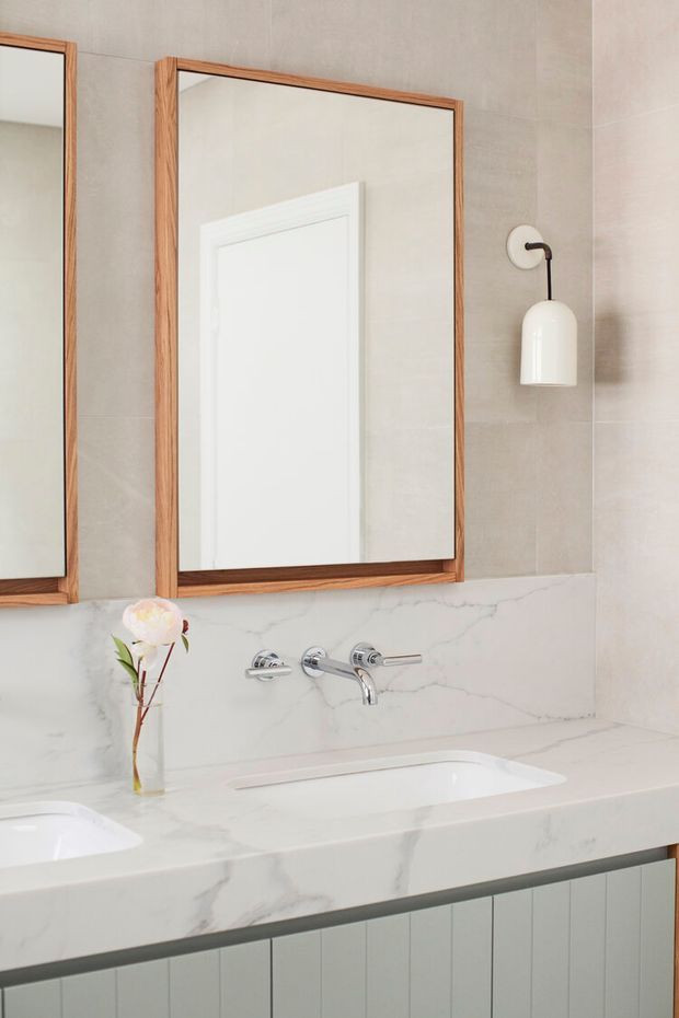 A refined contemporary bathroom vanity featuring a double sink, white veined stone benchtop and splashback, pale grey tiled walls, vertical panelled cabinetry, timber-framed mirrors, and a minimalist wall-mounted pendant light. Chrome wall-mounted tapware and a small vase with flowers sit on the vanity.