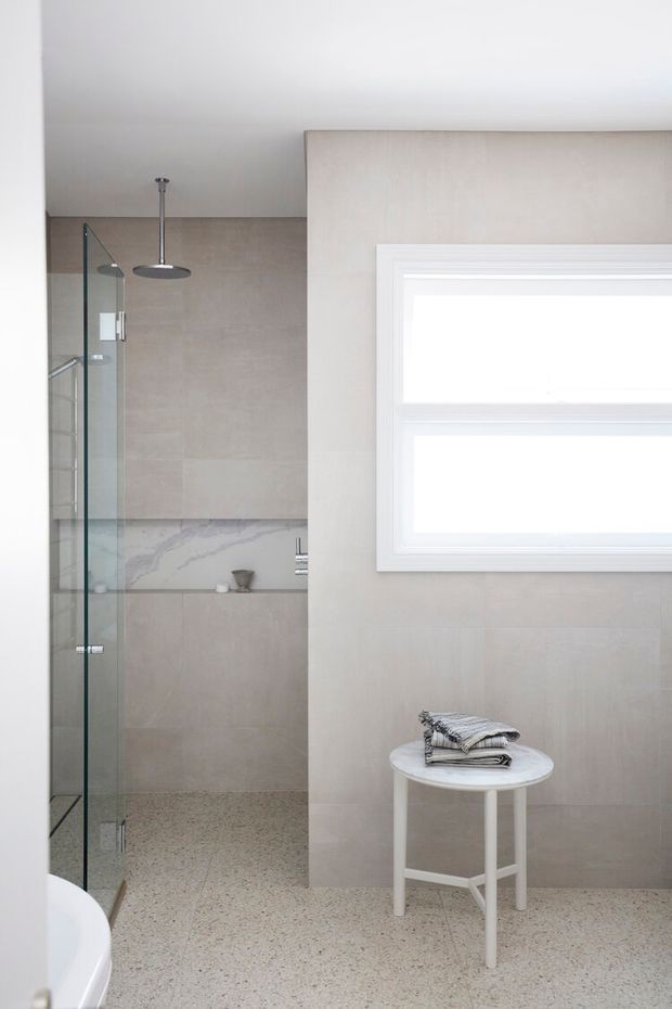 Bright, refined contemporary bathroom featuring a walk-in glass shower, large-format pale stone-look wall tiles, a marble-look recessed shelf, terrazzo-style flooring, and a high horizontal white-framed window. A small white stool with folded towels adds a soft furnishing accent.