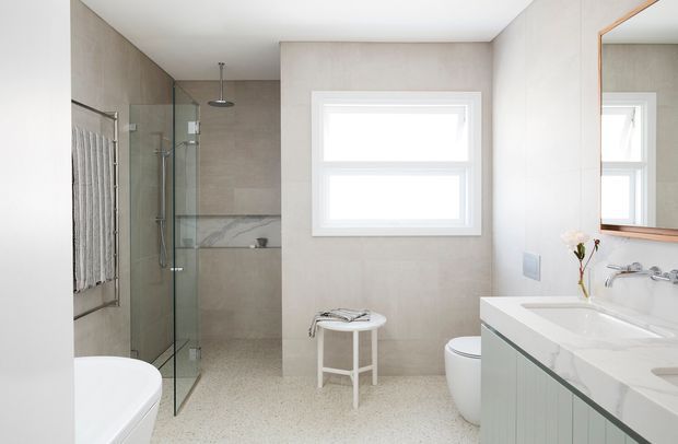 Bright contemporary bathroom with a walk-in glass shower, freestanding bathtub, toilet, floating vanity, large wall mirror, and a white-framed window. The room features light beige wall tiling, pale terrazzo-style flooring, and clean white fixtures.