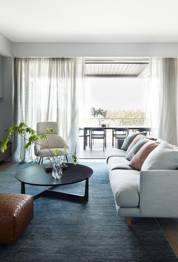 Bright, refined living room with a large pale sectional sofa, rounded armchair, dark oval coffee table, woven leather ottoman, and blue-grey rug. Full-height sheer curtains frame an open dining area and covered balcony, with abundant natural light and soft neutral furnishings.