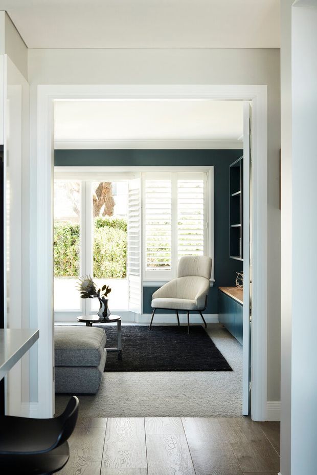 A refined sitting area viewed through a wide doorway, with dark teal built-in joinery, white plantation shutters, a cream upholstered lounge chair, grey sofa, black coffee table and rug. The room is bright and calm, with pale timber flooring in the foreground and a neutral painted interior.