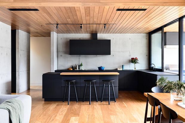 Open-plan contemporary kitchen with a large matte-black island and cabinetry, three bar stools, black appliances and fixtures, a concrete feature wall, warm timber flooring and a timber-lined ceiling. Full-height black-framed glazing and a nearby dining area bring in abundant natural light.