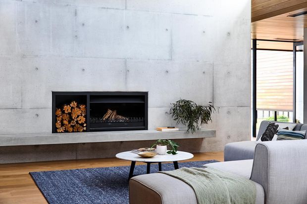 Contemporary living room with a smooth exposed concrete feature wall incorporating a built-in fireplace and firewood niche. A long concrete hearth shelf, low round coffee table, upholstered sectional seating, blue area rug, timber flooring, timber ceiling and expansive glazing create a refined, warm modern interior.