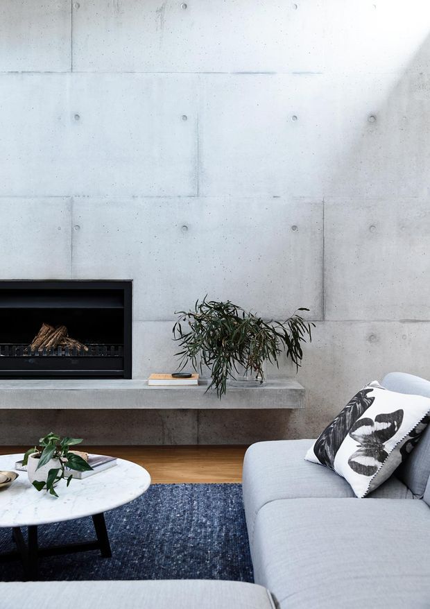 Contemporary living room featuring a poured concrete feature wall and built-in concrete fireplace hearth, light grey sectional sofa, marble coffee table, dark blue rug, and indoor greenery. The restrained palette and exposed concrete create a calm, minimalist atmosphere.