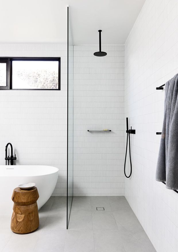Bright contemporary bathroom with a walk-in shower enclosed by a slim black-framed glass screen, white vertically lined wall tiles, pale grey floor tiles, a freestanding white bathtub, black shower and tapware, and a sculptural timber stool. A high horizontal window provides natural light.