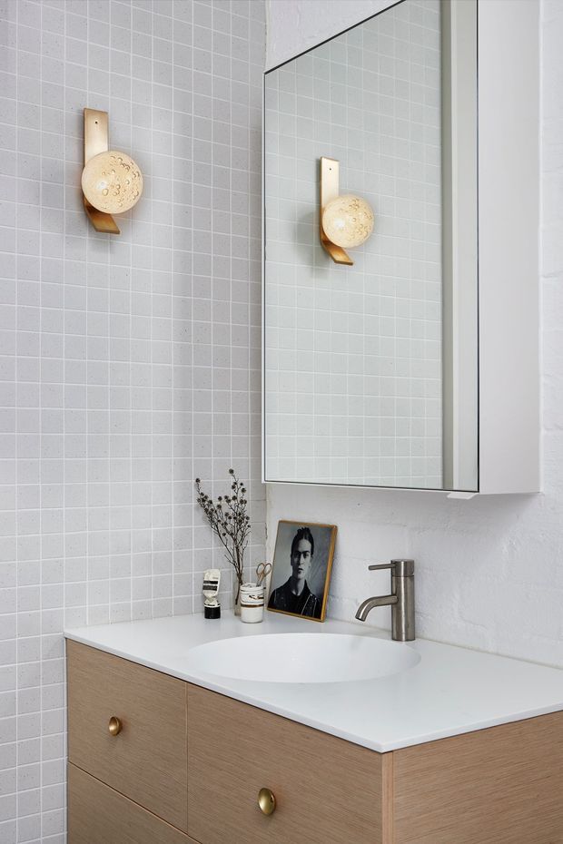 A compact, contemporary bathroom vanity with a white integrated basin, light timber-grain cabinetry and brass-toned knobs. Pale small-format square tiles cover the wall behind the vanity, while a large mirrored cabinet reflects the wall-mounted decorative light. A brushed metal faucet, framed portrait and small accessories sit on the white countertop.