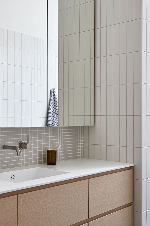 Bright, minimalist bathroom vanity with a white integrated basin, wall-mounted brushed metal tapware, pale wood-look cabinetry, white tiled walls, and a small grey mosaic tile splashback. A large mirrored cabinet spans the upper wall.