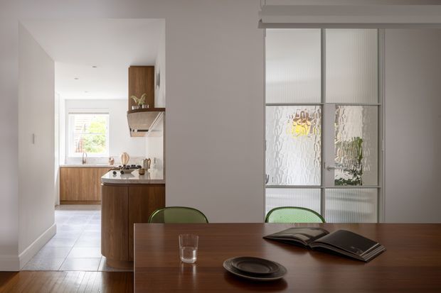A refined open-plan dining area adjoining a compact kitchen. The foreground features a dark timber dining table with simple tableware and books, while the kitchen has warm timber cabinetry, a pale benchtop, stainless-steel appliances, and light tiled flooring. A translucent fluted-glass partition with slim framing separates the dining area from another space, and soft natural light enters through the kitchen window.