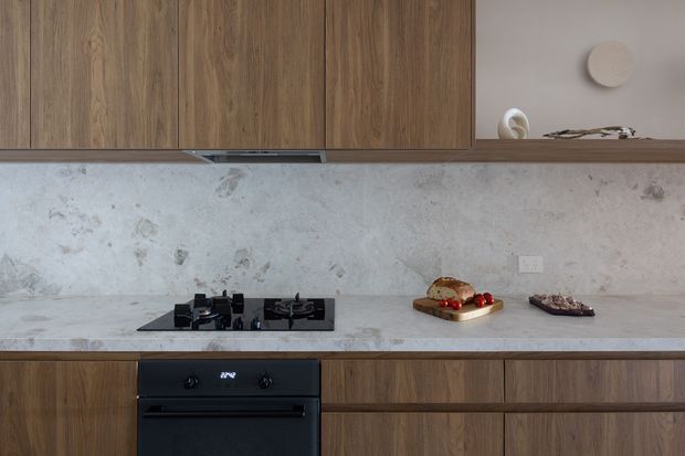A close-up view of a contemporary kitchen featuring full-height warm timber cabinetry, a pale grey mottled stone backsplash and benchtop, and integrated black cooking appliances. A gas cooktop and oven sit within the lower cabinetry, while a slim open shelf and minimal decorative objects add subtle detail. The composition is clean and understated with natural material tones.