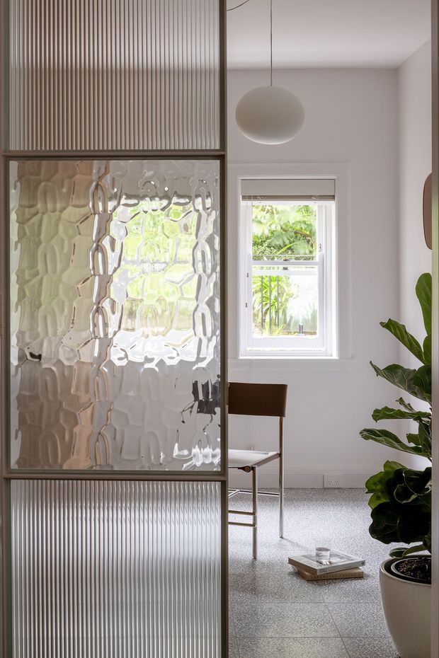 A bright, refined interior vignette featuring a full-height framed glass partition or sliding screen. The partition combines vertically ribbed translucent glass panels with a central textured privacy-glass panel in slim dark frames. Behind it is a minimalist dining or living area with a white-framed window, a simple dark wood chair, a spherical pendant light, pale walls, a terrazzo-like floor, and a large leafy indoor plant.