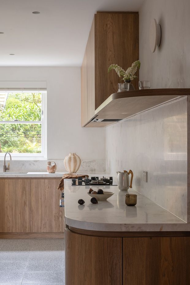 A warm, refined contemporary kitchen featuring rich timber cabinetry, a pale stone benchtop with a rounded peninsula edge, integrated stainless-steel cooking appliances, and a light tiled or stone splashback. A window above the sink brings in soft natural light, while understated decor and recessed lighting create a calm, elegant atmosphere.
