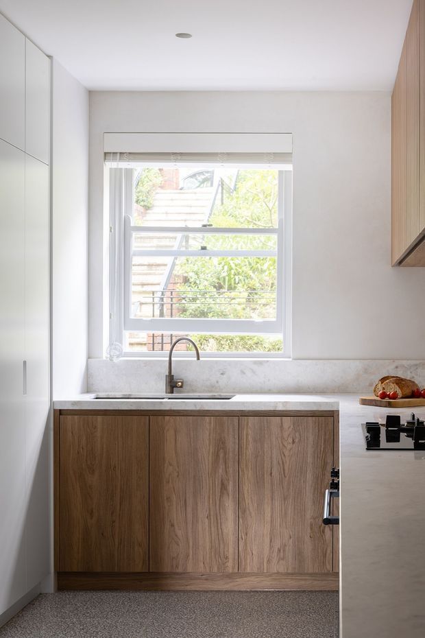 A bright, compact contemporary kitchen focused on a timber-fronted base cabinet run beneath a window. The kitchen features a pale stone benchtop and integrated sink with a brushed metal tap, white full-height cabinetry on the left, and a partially visible pale island or adjoining counter on the right with a black cooktop. Natural light enters through the large window, creating a clean, warm, understated interior.