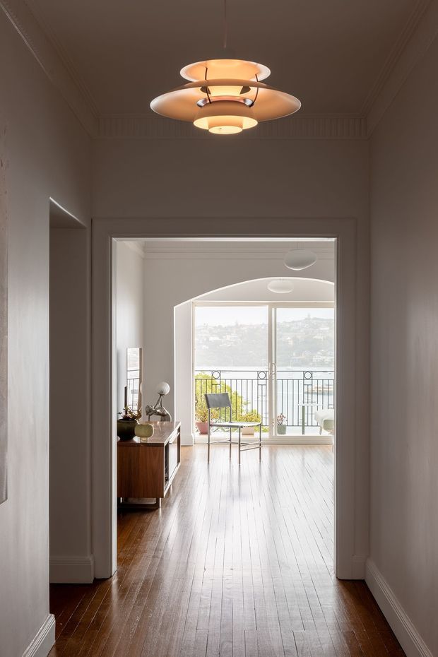 A refined interior view looking through a wide doorway from a narrow hallway into a bright living area. The hallway has white painted walls, ornate ceiling cornices and moulding, and polished dark timber flooring. A sculptural layered pendant light hangs centrally overhead. Beyond the doorway, the living space features pale timber flooring, curved ceiling detailing, a mid-century timber sideboard, minimalist furnishings, and full-height glazing opening to a balcony with a visible water and hillside outlook.