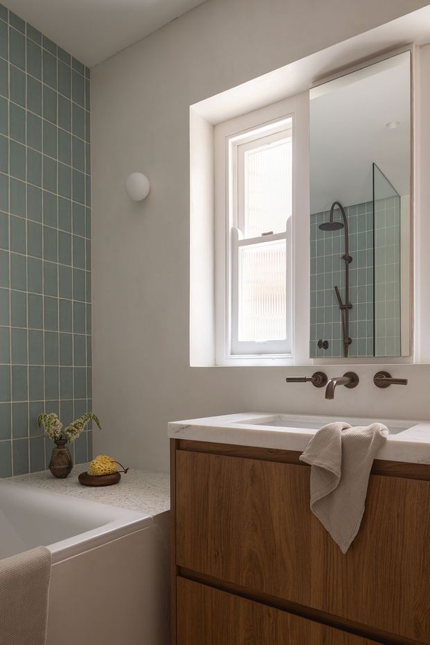 A softly lit contemporary bathroom with a white bathtub, wall-mounted timber vanity, pale stone benchtop, integrated basin, wall-mounted bronze tapware, and a large mirrored cabinet. Muted blue-green rectangular tiles form the shower and bath surround, with a frosted window providing natural light. Neutral painted walls, simple rounded wall lighting, and restrained styling create a calm, refined atmosphere.