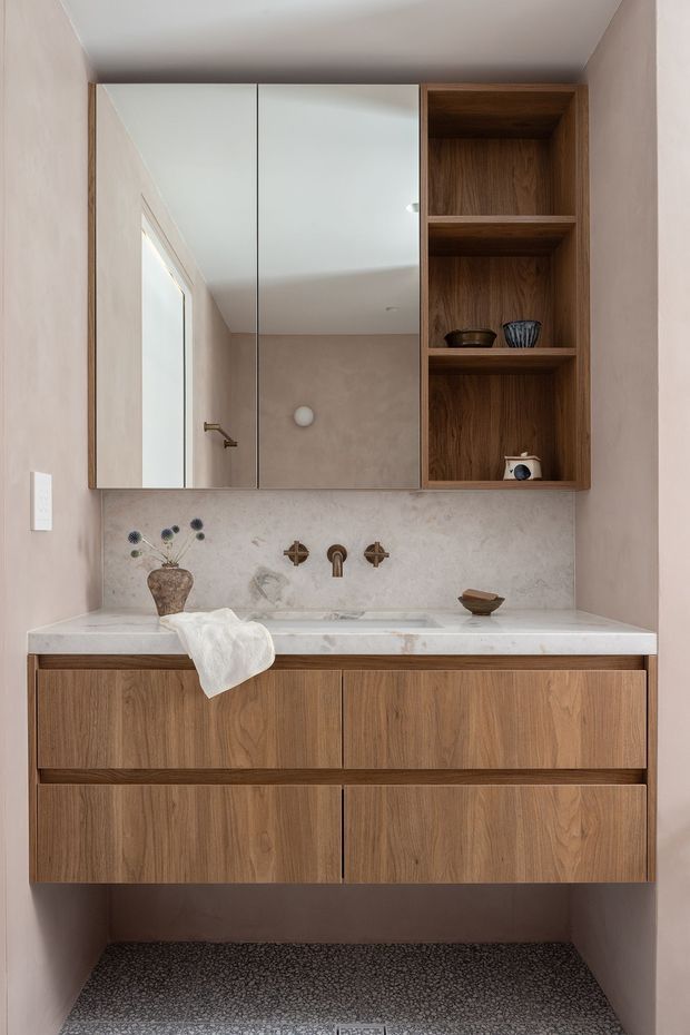 A compact, refined bathroom vanity area with a floating natural wood cabinet, white stone countertop and integrated basin, wall-mounted bronze-toned tapware, mirrored upper cabinetry, and open wood shelving. The walls are finished in a warm blush-beige tone, with a mottled stone backsplash and small dark pebble-style flooring. Minimal decorative ceramics and a vase add restrained styling.