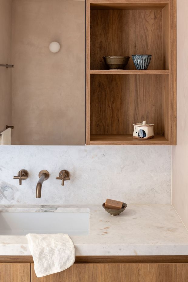 Close-up view of a refined bathroom vanity featuring a pale stone or plaster-look benchtop and integrated rectangular basin, bronze wall-mounted tapware, warm timber cabinetry, and an open timber display niche with ceramic vessels. The walls and countertop have a softly mottled, natural finish, creating a warm, tactile, contemporary atmosphere.