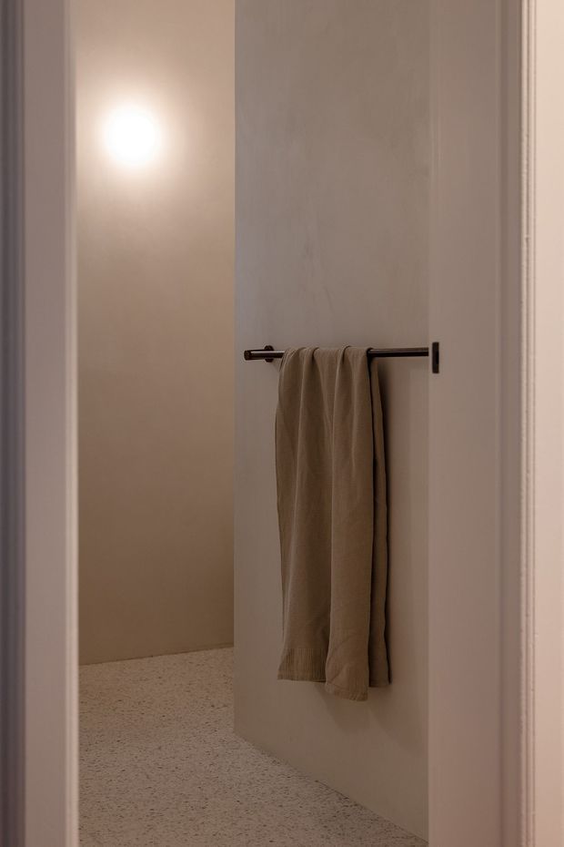 A softly lit minimalist interior detail showing a warm beige towel hanging from a dark metal wall-mounted rail on a pale plaster wall. A doorway or opening reveals a softly illuminated adjacent area, and the floor appears to be light terrazzo or fine-speckled concrete. The composition is quiet, restrained, and highly minimal.