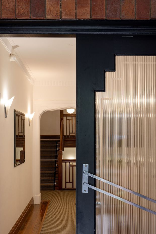 View through a partially open black-framed door into a refined heritage-style hallway and stair landing. The space features polished timber flooring, a carpet runner, cream painted walls with decorative cornices and moulded architraves, traditional wall sconces, timber stair balustrades, and a distinctive ribbed translucent glass door panel with a geometric stepped edge.