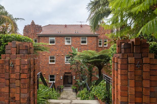 Exterior view of a two-storey red-brick residential building seen through a landscaped entry passage. The building has a pitched terracotta-tiled roof, brick chimney, regularly spaced white-framed windows, and a central recessed entrance reached by a paved stairway. Brick gate pillars and dense tropical planting frame the foreground.