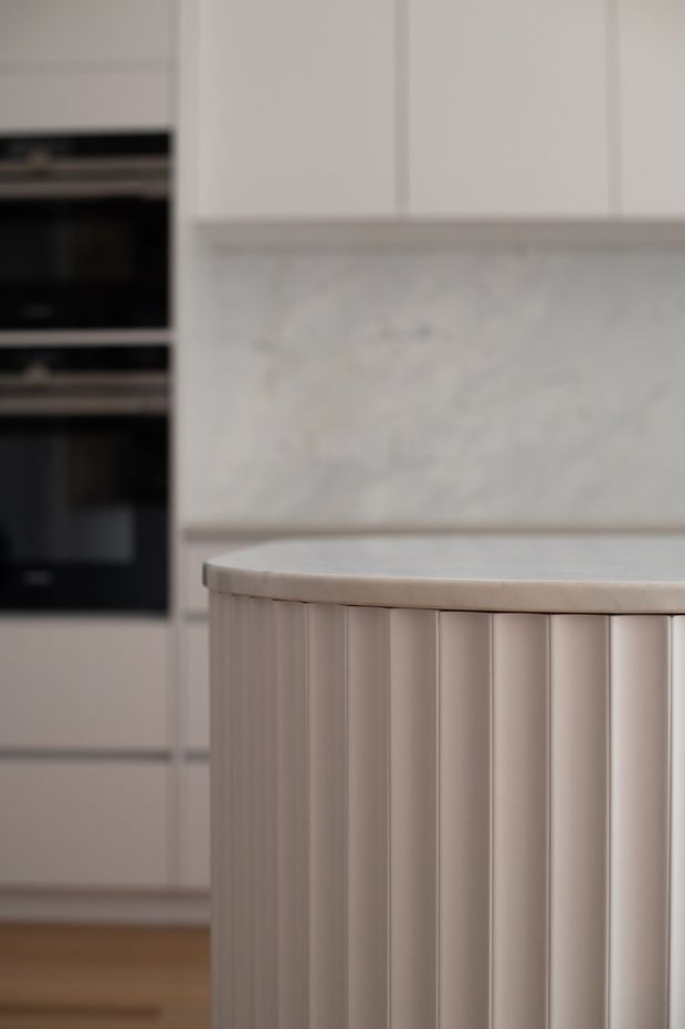 Close-up detail of a refined light-toned kitchen, featuring a curved island with vertical fluted paneling, a pale stone benchtop and matching stone splashback. White cabinetry and integrated black ovens are visible in the background, with timber flooring below.
