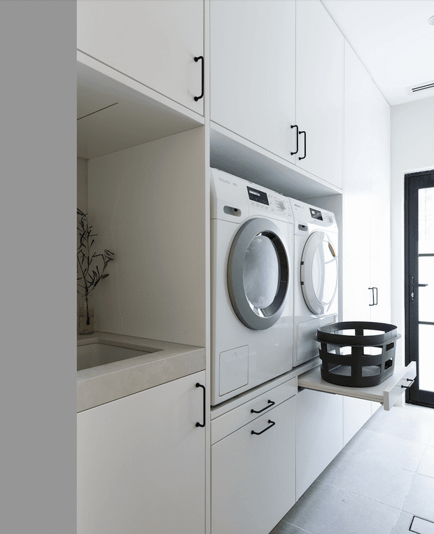 Bright, compact laundry room with stacked front-loading washing machine and dryer integrated into white built-in cabinetry. The room features black metal handles, pale tiled flooring, a deep utility sink or basin at left, and a black-framed glazed door on the right. Clean, functional contemporary fit-out with strong white and grey tones.