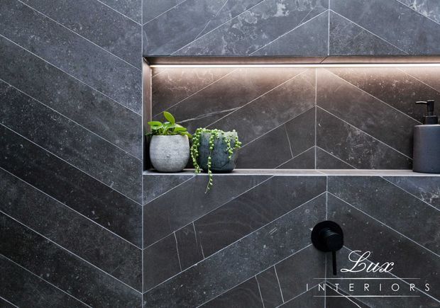 Close-up of a dark, luxurious tiled bathroom wall featuring a geometric chevron layout, a recessed illuminated shelf with potted plants and a soap dispenser, and a black wall-mounted tap or control. The tiles have a charcoal stone-like finish with contrasting grout lines.
