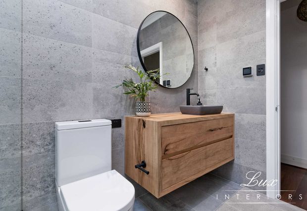 Modern bathroom featuring large-format grey concrete-look wall tiles, dark grey floor tiles, a wall-mounted natural timber vanity with integrated drawers, a vessel basin, matte black tapware, round black-framed mirror, and a white toilet. A doorway reveals a small glimpse of an adjacent room.