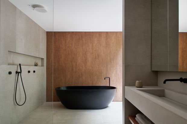 Contemporary minimalist bathroom featuring a freestanding matte black oval bathtub, wall-mounted black tapware and shower fittings, pale concrete-look wall and floor surfaces, warm timber wall panelling, and a floating light-coloured vanity with integrated basin. A frameless glass shower screen divides the wet area, creating a calm, refined space with strong material contrast.