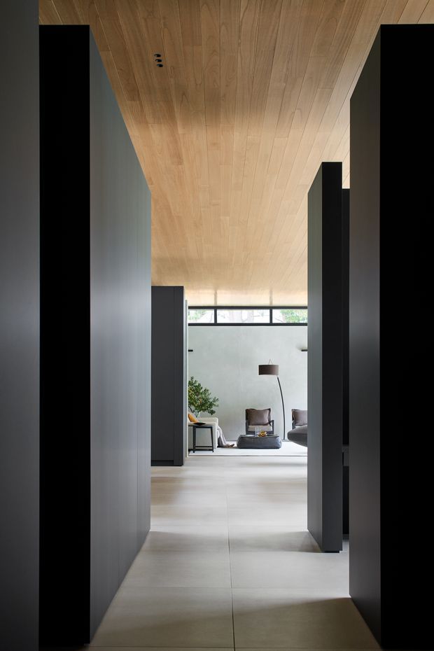 A minimalist interior corridor leading into a refined living room. Tall dark built-in wall planes frame the passage, while a continuous light timber-lined ceiling extends toward the bright seating area. Large pale floor tiles, a narrow clerestory window, sculptural floor lamp, armchairs, and low coffee table create a calm, contemporary architectural composition.