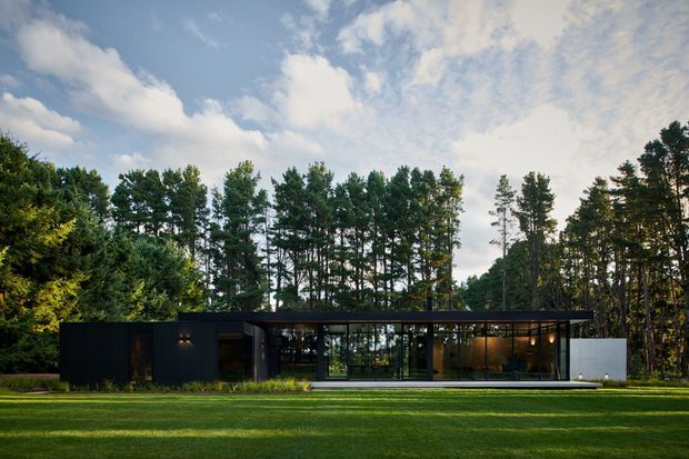 A low, single-storey modern house set within a wooded landscape. The building has a long, rectilinear form with dark opaque wall cladding, extensive floor-to-ceiling glazing, and a thin flat roof. It is viewed from a broad manicured lawn, with tall trees forming a dense backdrop under a partly cloudy sky.
