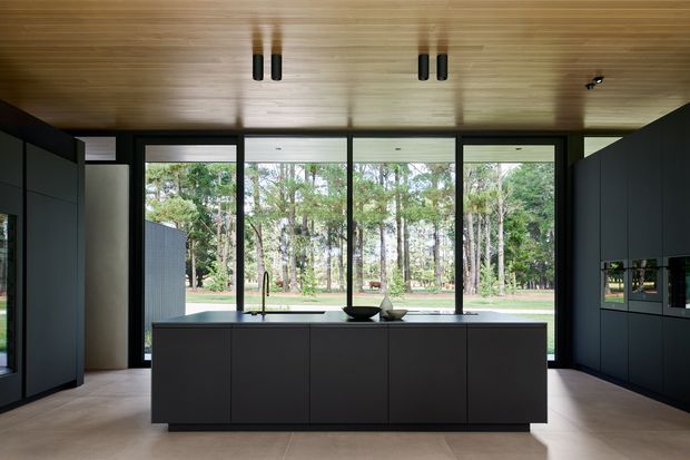A refined contemporary kitchen with a large dark island, extensive floor-to-ceiling glazing overlooking a wooded garden, dark built-in cabinetry on both sides, and a warm timber-lined ceiling with linear integrated lighting.