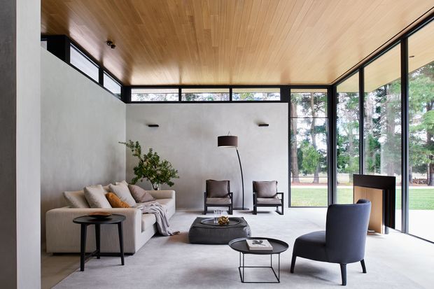 A refined contemporary living room with full-height black-framed glazing overlooking a landscaped garden. The room features a pale upholstered sofa, sculptural lounge chairs, low black coffee tables, a freestanding floor lamp, smooth light walls, a warm timber-lined ceiling, and a soft neutral carpet.